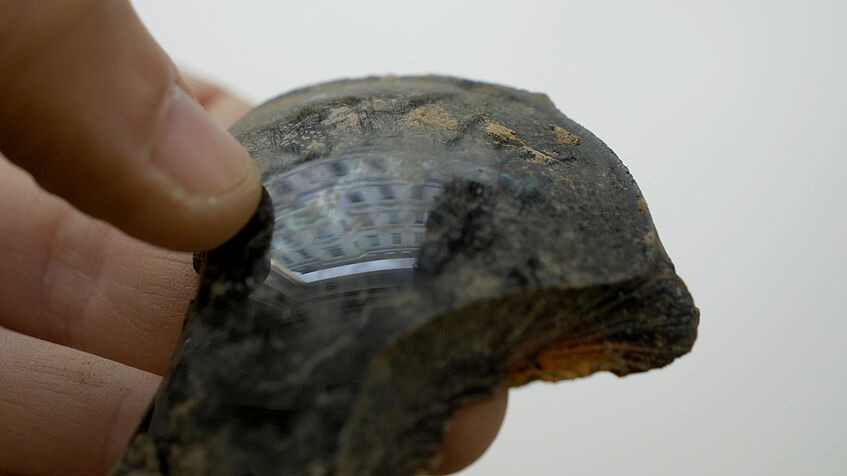 Close-up of an archaeological find - a shiny piece of glass or plastic, reflecting houses in the background